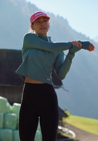 Žena v sportovním oblečení a růžové kšiltovce se protahuje venku, s horami a balíky sena v pozadí.