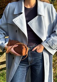 Jasnoniebieski płaszcz trencz z teksturowanej tkaniny, zestawiony z ciemnymi jeansami oraz małą brązową zamszową saszetką z zamkiem i paskiem.