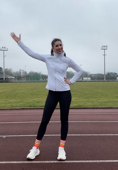 Chaqueta blanca con cremallera, leggings negros y calcetines a rayas de colores. De pie en una pista, saludando con un brazo. Campo de hierba de fondo.