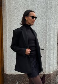Blazer mare de culoare neagră peste un guler roulé negru, asortat cu o fustă mini texturată de culoare neagră. Accesoriile includ ochelari de soare negri și cercei aurii.