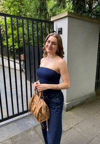 Modra brezramna majica, temno modri široki kavbojki in svetlo rjava torba z zadrgo in detajli. Ozadje prikazuje črno ograjo in zelenje.