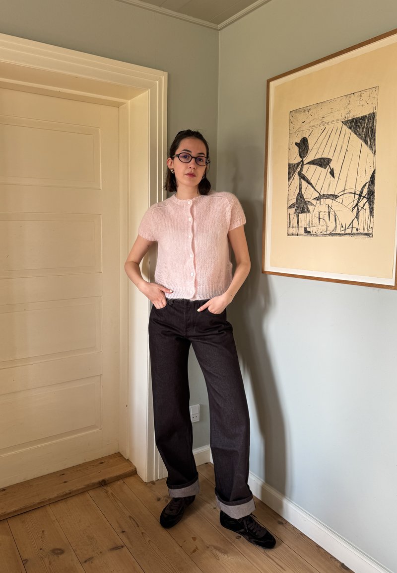 Suéter de malha cortado em rosa claro com mangas curtas e fecho de botão, combinado com jeans largos escuros, com punhos dobrados e sapatos pretos.