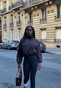 Kvinne med langt svart hår går på en gate i byen iført en brun jakke, svarte leggings og hvite sko, mens hun bærer en mørkebrun håndveske.