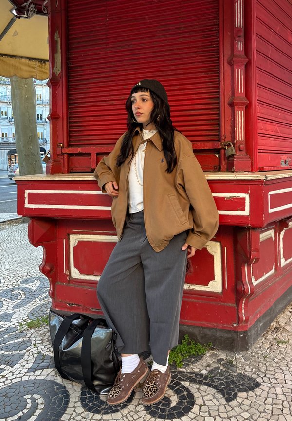 Nuori nainen, jolla on ruskea takki, harmaat housut ja leopardikuviolliset kengät, nojaa punaiseen puiseen kojueen kuviollisella jalkakäytävällä.