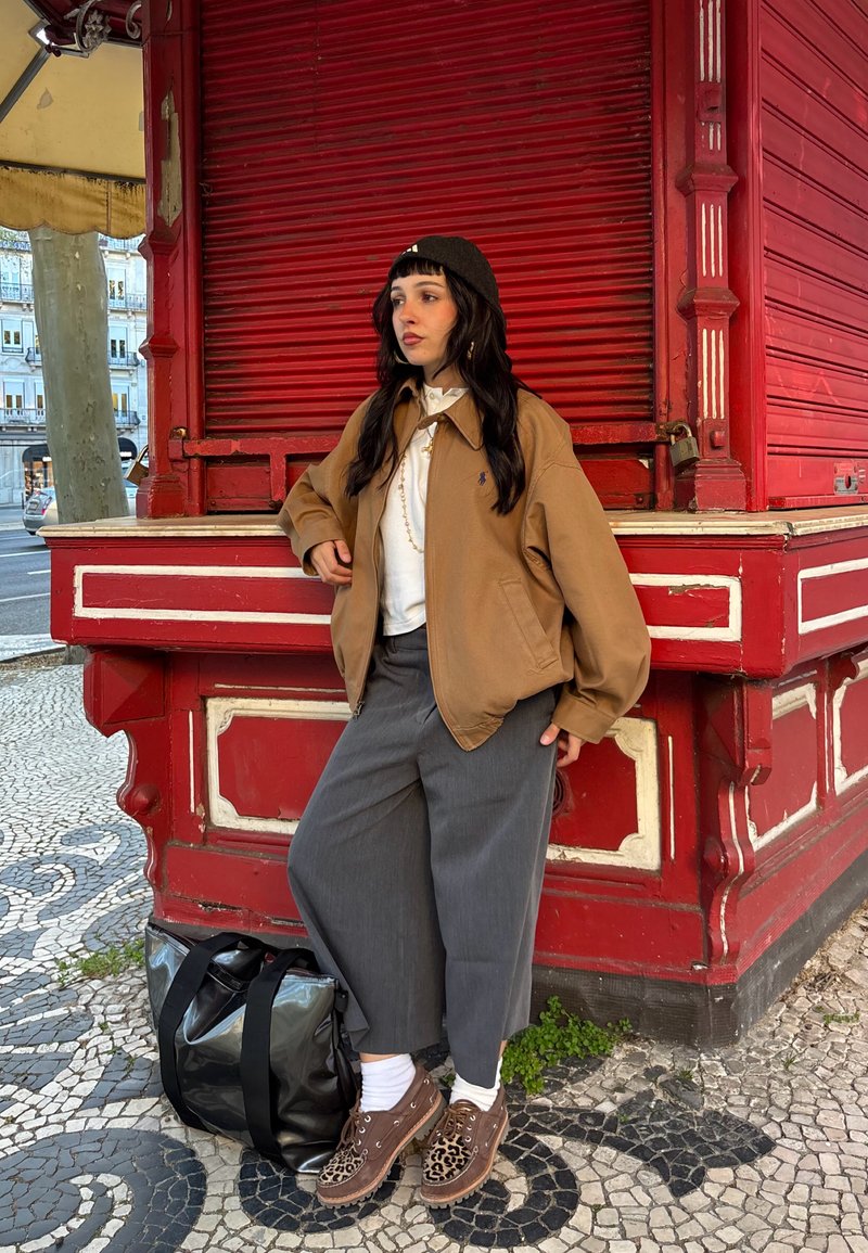 Mladá žena v hnědé bundě, šedých kalhotách a botách s leopardím vzorem se opírá o červený dřevěný stánek na vzorované chodníkové dlažbě.