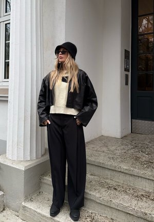 Mujer con sombrero tipo bucket negro, gafas de sol, chaqueta de cuero negra recortada, suéter crema y pantalones negros anchos, de pie sobre escalones de piedra junto a una columna blanca.