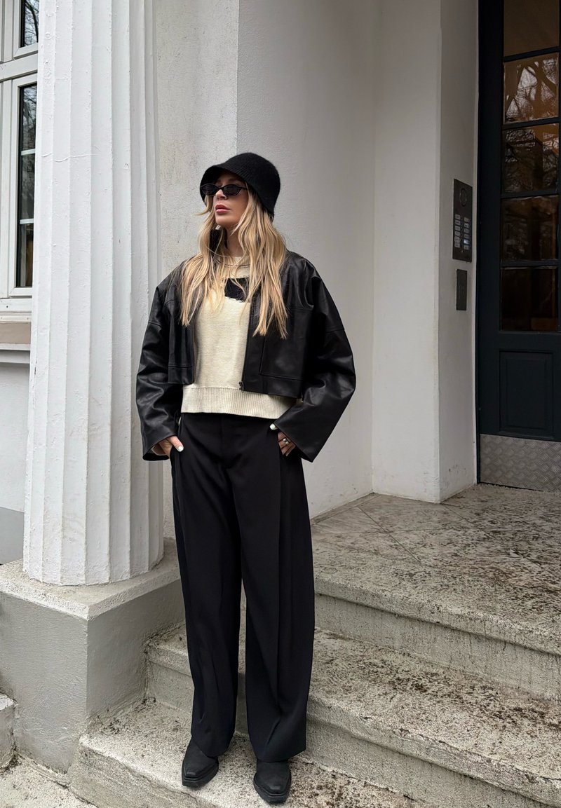 Femme portant un chapeau seau noir, des lunettes de soleil, une veste en cuir noire courte, un pull crème et un pantalon large noir debout sur des marches en pierre près d'une colonne blanche.