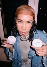 Moira cosmetics, een doorzichtige pot met roze crème en een witte pot met blauwe tekst. Lang krullend haar en een spijkerjack, vastgehouden in beide handen.