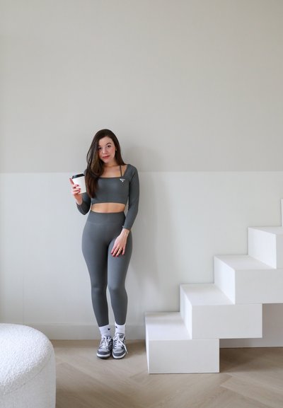 Top corto de manga larga gris con textura de canalé y leggings, combinados con zapatillas blancas y calcetines hasta el tobillo, sosteniendo una taza de café mientras está de pie junto a unas escaleras blancas.