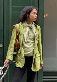 Groene, gestructureerde blazer met een getailleerde snit, gedragen over een lichtgroene top. Paar zwarte leggings en een bruine handtas. Gepatternde accessoire in de hand.