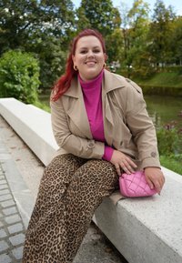 Μπεζ παλτό πάνω από ροζ πουλόβερ, συνδυασμένο με παντελόνι με λεοπάρ σχέδιο. Κρατώντας μια ροζ τσάντα με καπιτονέ σχέδιο. Εξωτερικός χώρος με πράσινη βλάστηση.