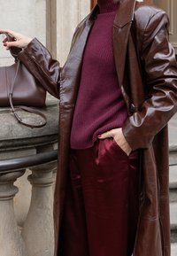 Palton lung din piele maronie, pulover din lână bordou, pantaloni din satin burgundy, poșetă asortată, mâinile în buzunare, texturi netede, croi ajustat.