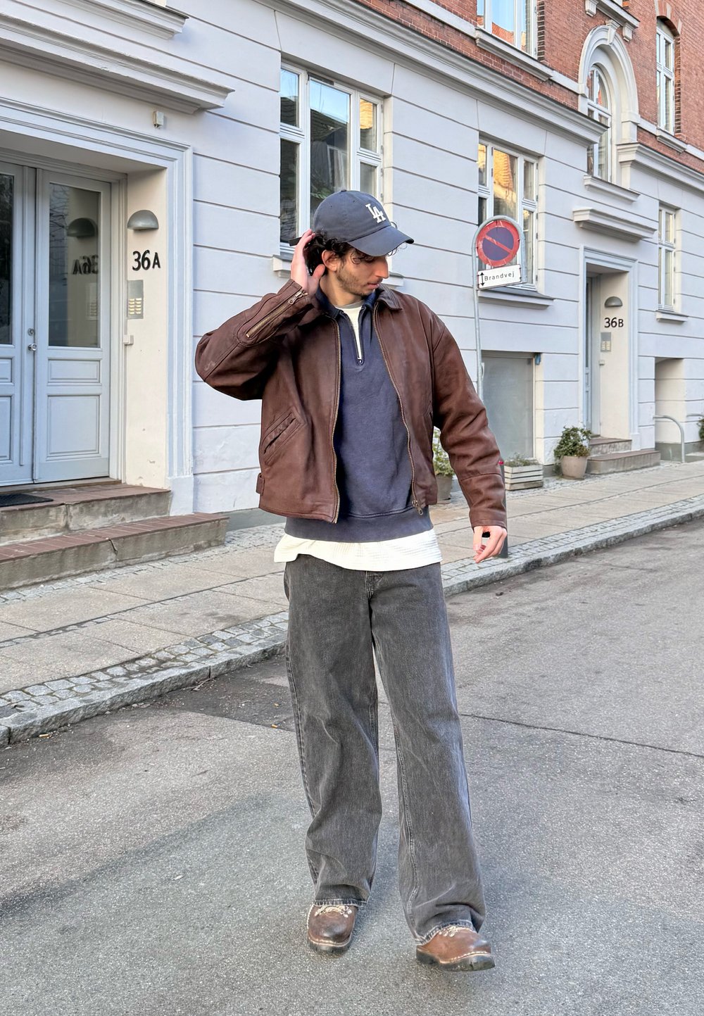 Homme portant une veste en cuir marron, une casquette grise et un jean gris ample, debout dans une rue de la ville près de bâtiments blancs et en briques.