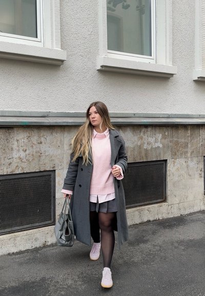 Joven caminando por la acera con abrigo gris, suéter rosa, falda negra y llevando un bolso gris en un entorno urbano.
