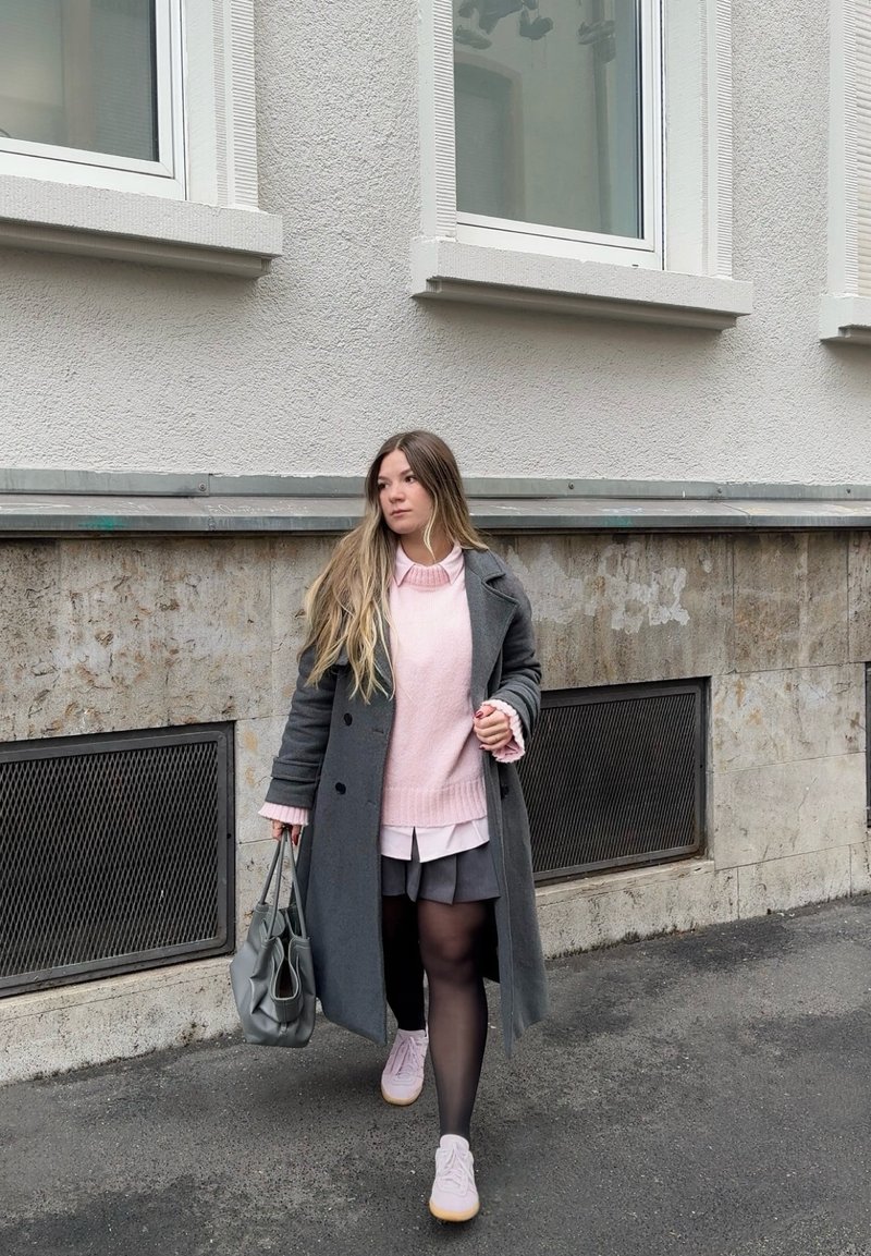 Jeune femme marchant sur le trottoir, portant un manteau gris, un pull rose, une jupe noire, et portant un sac à main gris dans un cadre urbain.