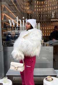 Femme portant une veste blanche en fourrure et un bonnet, tenant un sac à main beige, debout devant un café avec une fenêtre en verre réfléchissant et une terrasse avec des sièges.