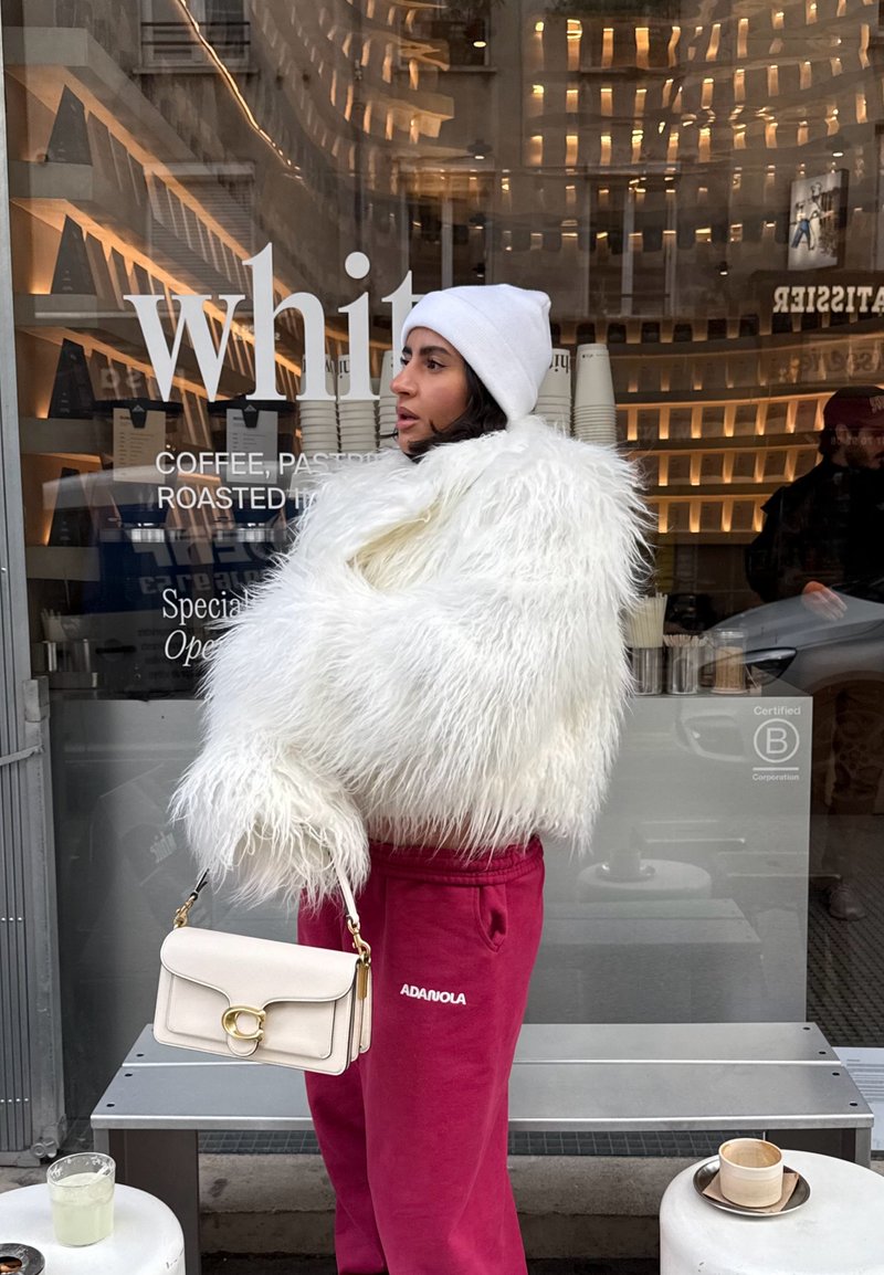 Woman in white furry jacket and beanie holding beige handbag, standing outside café with reflective glass window and seating area.