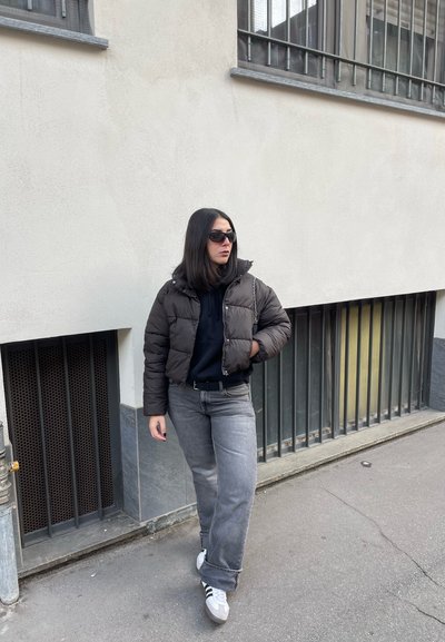 Chaqueta de plumas marrón con cuello alto, sudadera negra con capucha, jeans rectos grises y zapatillas blancas. Unas gafas de sol negras completan el look.