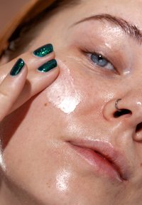 Hydraterende crème aangebracht op de wangen van een gezicht met sproeten. Groene glinsterende nagels en een zilveren oorring zijn zichtbaar. Gladde, glanzende textuur.