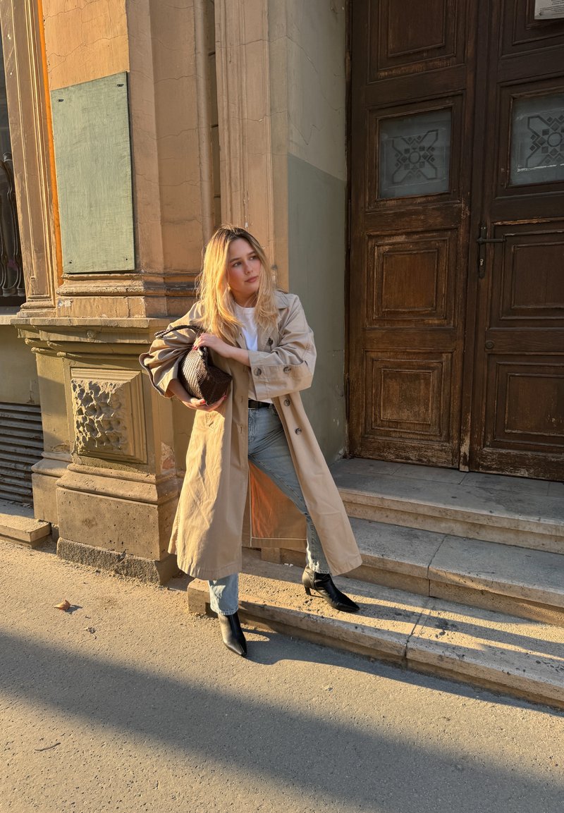 Jeune femme en trench beige, jean clair et bottes noires ajuste son sac en se tenant sur des marches en pierre devant un immeuble.