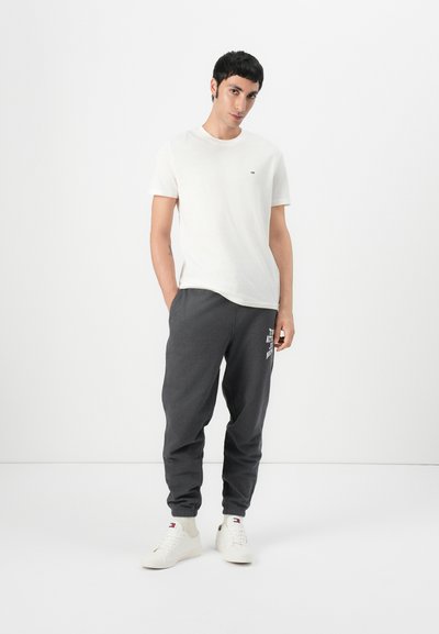 Camiseta blanca de algodón con un pequeño logo en el pecho, combinada con pantalones de chándal gris oscuro que tienen texto impreso, y zapatillas blancas.