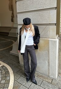 Zwarte blazer, wit tanktopje, grijze wijde broek, zwarte baret en pluizige witte sjaal. Zwarte puntige hakken. Buitenomgeving met een gestructureerde muur.