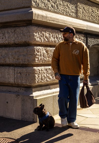 Jersey texturizado naranja con cuello de cremallera, vaqueros azules, zapatillas blancas y bolso marrón. Un bulldog francés negro con un arnés azul está sentado cerca.