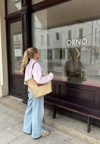 Gehaakte stro tas met bruine leren accenten, gecombineerd met een lichtroze gestreept shirt en wijde blauwe jeans. Vensterreflectie zichtbaar.