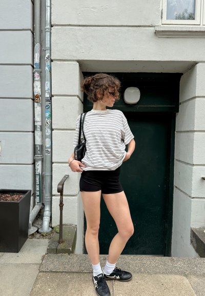 Camiseta de manga corta a rayas en blanco roto y negro, combinada con pantalones cortos negros. Zapatillas negras y calcetas blancas completan el atuendo.