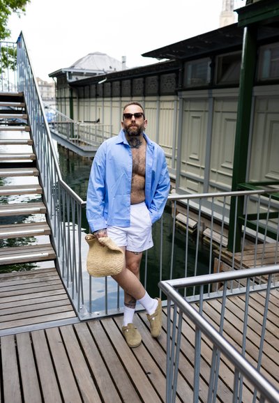 Camisa de botones azul claro, pantalones cortos blancos, zapatos beige, usando gafas de sol, sosteniendo una bolsa de mimbre, de pie en una terraza de madera junto al agua.