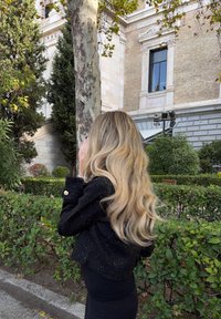 Giacca nera testurizzata con accenti dorati, lunghi capelli biondi ondulati, in piedi contro un fondo di vegetazione e architettura in pietra.