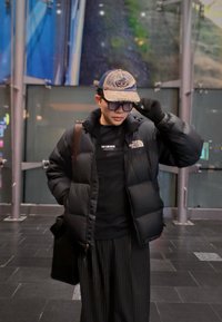 Čierna prešívaná bunda s logom, čierne tričko, pruhované nohavice a maskáčová čiapka. Osoba drží rukavicu, stojí v interiéri pri sklenených dverách.