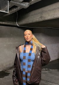 Brun läderoversizedjacka, blå stickad tröja och en scarf med blå och bruna rutmönster. Modellen står i en industriell miljö med grå väggar.