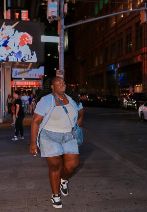 Camisa azul clara de botones sobre una camiseta blanca sin mangas, pantalones cortos de mezclilla, zapatillas negras y blancas, y un pequeño bolso cruzado azul. Escena urbana nocturna.