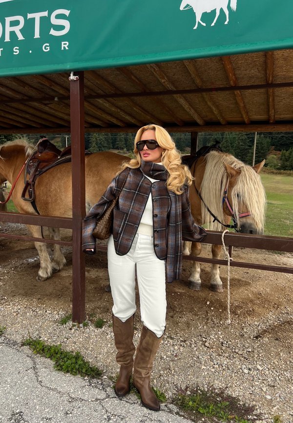 Mujer con chaqueta marrón de cuadros, pantalones blancos y botas altas, de pie junto a dos caballos ensillados bajo un refugio verde en un entorno rural al aire libre.