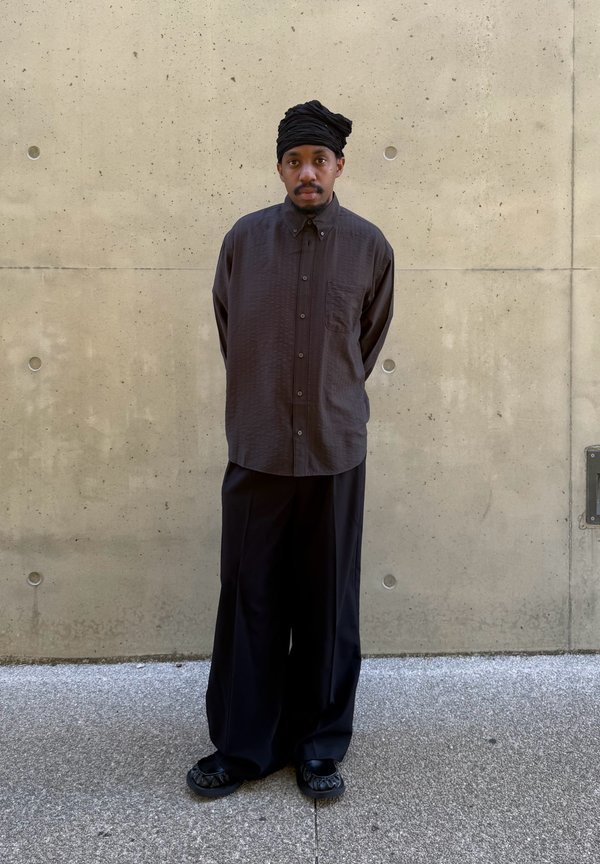 Homme portant un turban noir, une chemise boutonnée oversize foncée et un pantalon large noir, debout les mains dans le dos contre un mur en béton.
