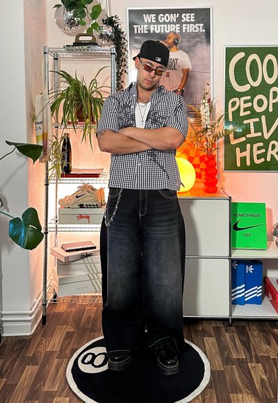 Camisa de cuadros en blanco y negro de manga corta, jeans negros holgados, cadena negra y gafas de sol. De pie sobre una alfombra redonda negra con un logotipo.