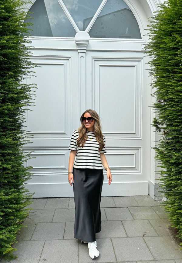 Top de manga corta a rayas blancas y negras, falda maxi de satén negra, zapatillas blancas, de pie frente a una puerta blanca con molduras decorativas.