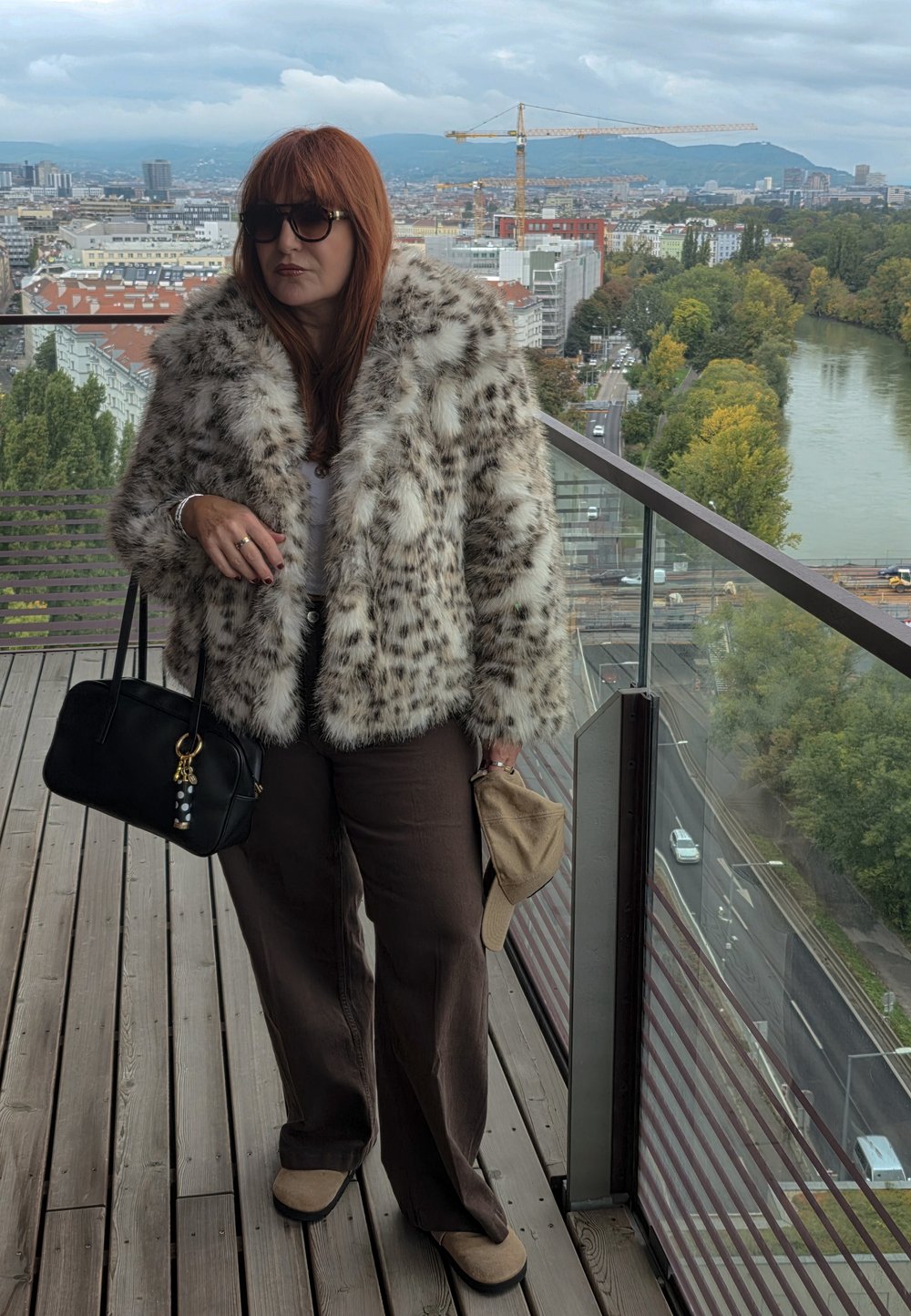 Leopard print faux fur coat, white top, brown wide-leg pants, black handbag with decorative charms, standing on a balcony with a city view.