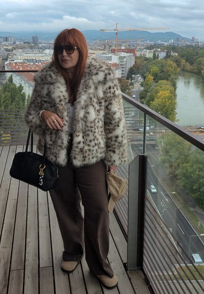 Abrigo de piel sintética con estampado de leopardo, top blanco, pantalones anchos marrones, bolso negro con adornos decorativos, de pie en un balcón con vistas a la ciudad.