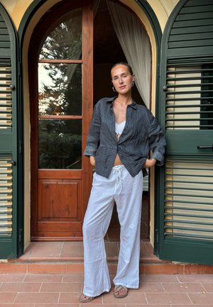 Camisa de botones negra a rayas, pantalones blancos texturizados, de pie frente a una puerta de madera con contraventanas verdes. Azulejos de piso en tonos tierra visibles.