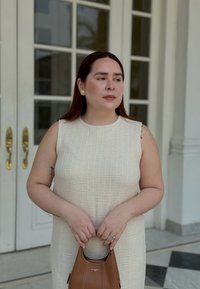 Robe sans manches de couleur claire et texturée avec un col rond, accompagnée d'un petit sac à main marron. Boucles d'oreilles en or et détail de tatouage visible.