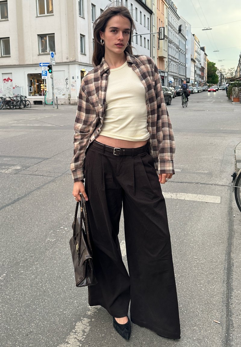 Jeune femme en crop top beige, chemise à carreaux, pantalon large noir et talons, tenant un sac à main marron dans une rue urbaine avec des bâtiments et un cycliste derrière.
