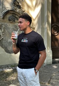 T-shirt noir à manches courtes avec un texte coloré qui dit "Société de 032c." Associé à un short blanc et un gobelet transparent avec une paille.