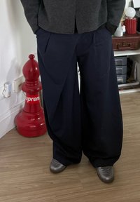 Marineblauwe broek met wijde pijpen en een geplooid ontwerp, gemaakt van een gestructureerde stof. Gecombineerd met grijze sneakers, staand op een houten vloer.