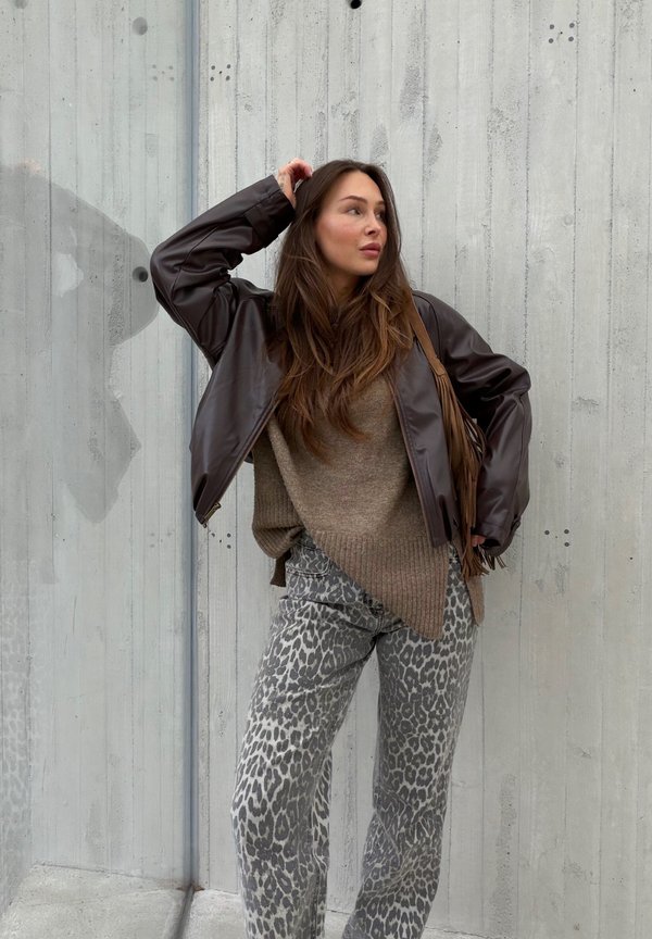 Femme aux longs cheveux bruns portant une veste en cuir marron, un pull beige et un pantalon gris à motif léopard, debout contre un mur en béton.