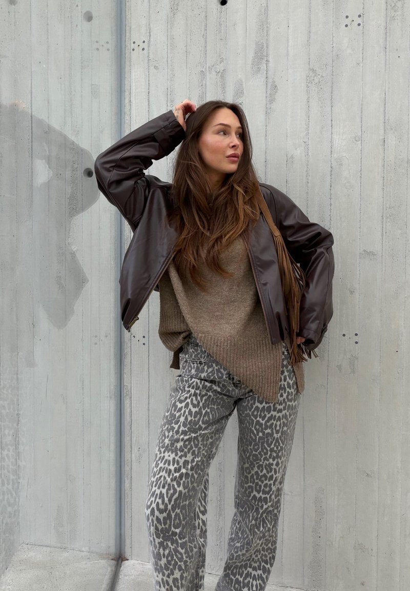 Woman with long brown hair wearing brown leather jacket, beige sweater, and gray leopard print pants stands against concrete wall.