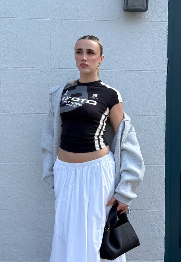 Giovane donna con top sportivo nero, gonna bianca e giacca grigia tiene una borsa nera, in piedi contro una parete e una porta chiara.