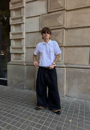 Camiseta polo blanca, pantalones vaqueros negros de gran tamaño, zapatos oscuros y accesorios. El entorno presenta una pared de piedra texturizada y un pavimento de baldosas.