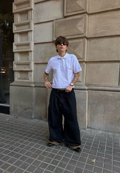 Camiseta polo blanca, pantalones vaqueros negros de gran tamaño, zapatos oscuros y accesorios. El entorno presenta una pared de piedra texturizada y un pavimento de baldosas.